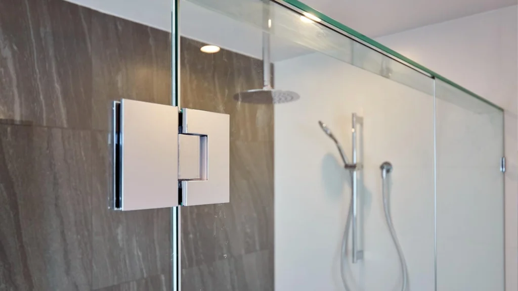 Heavy-duty chrome hinge mounted on a thick frameless tempered glass shower door in a modern hotel bathroom.