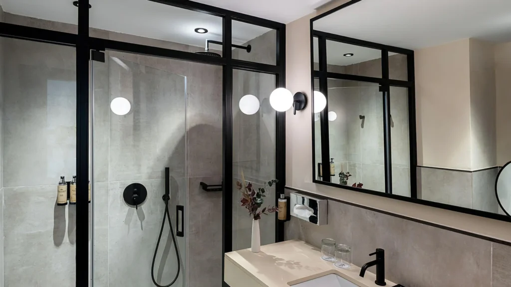 Floor-to-ceiling black framed glass shower enclosure custom fitted next to a hotel bathroom vanity layout.
