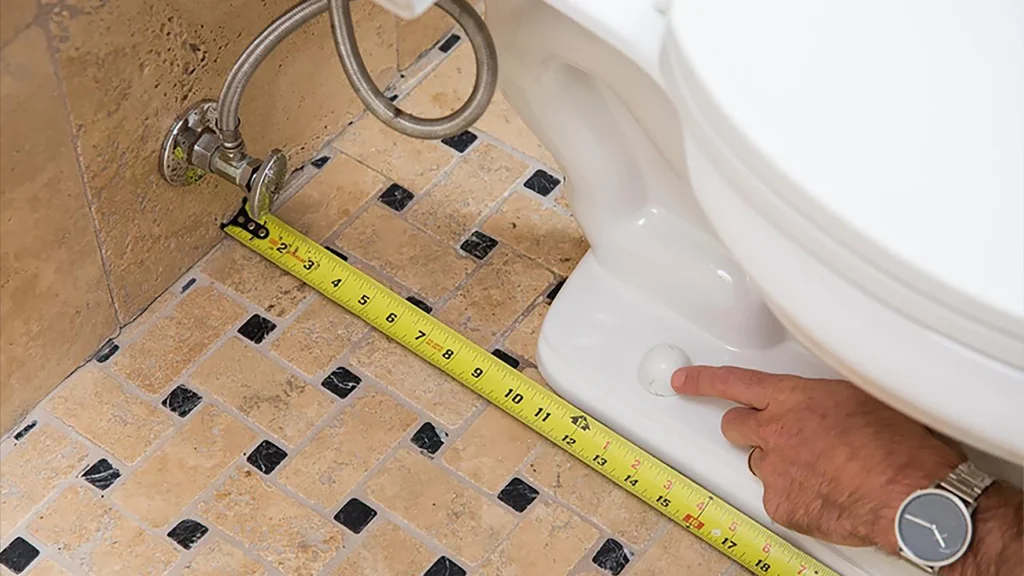 Close-up of measuring rough-in distance from the finished wall to the toilet bolt cap to verify exact clearance.