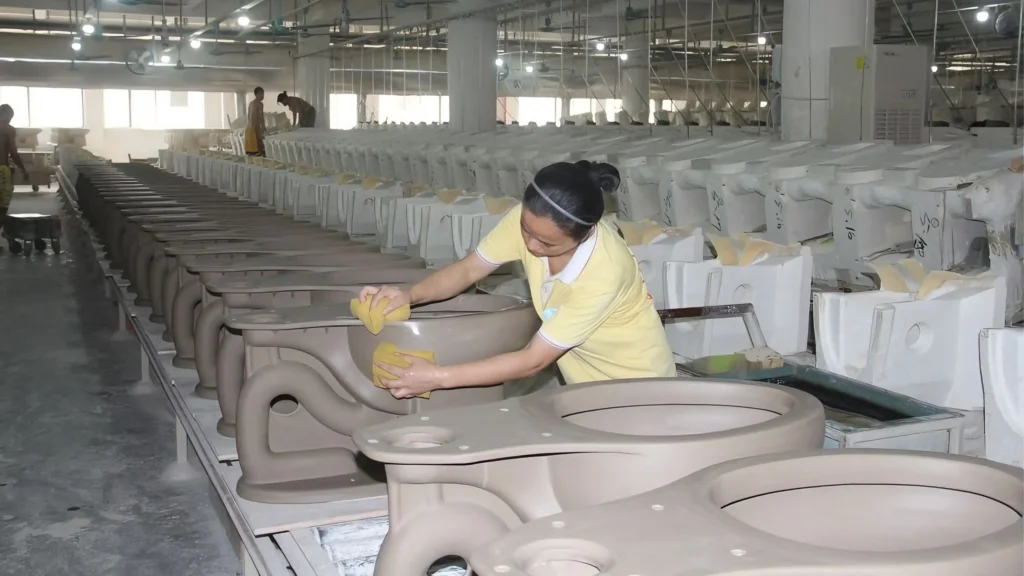 Factory worker hand-finishing ceramic toilet bodies on a production line for commercial project orders.
