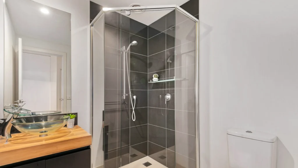 Corner pivot glass shower door with chrome hardware, fitting between vanity and toilet in a compact hotel bathroom.
