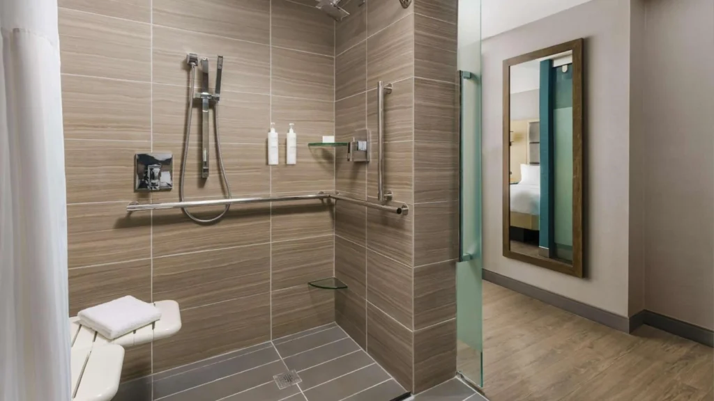 ADA roll-in shower with stainless steel grab bars, fold-down seat, and zero-barrier entry in a hotel bathroom.