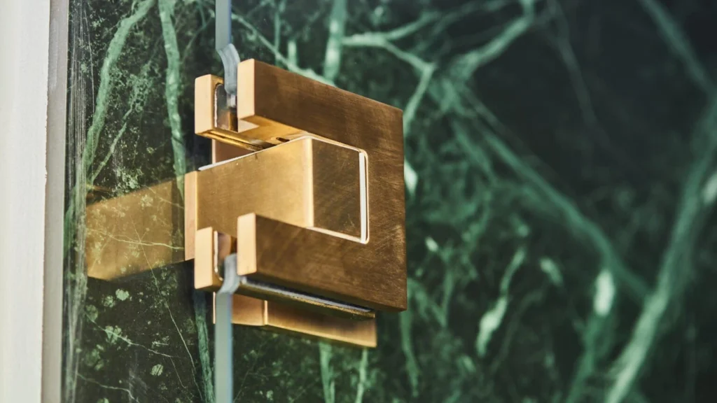 Brushed brass commercial shower door hinge mounted on frameless tempered glass in a luxury hotel bathroom.