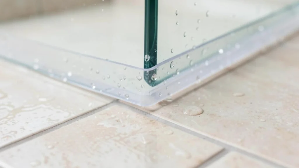 Clear plastic sweep seal on a frameless glass shower door with water droplets on a hotel bathroom curb.