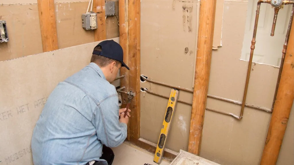 Plumber works on copper rough-in plumbing in an apartment wet area to verify dimensions before a bulk order, with a level nearby.